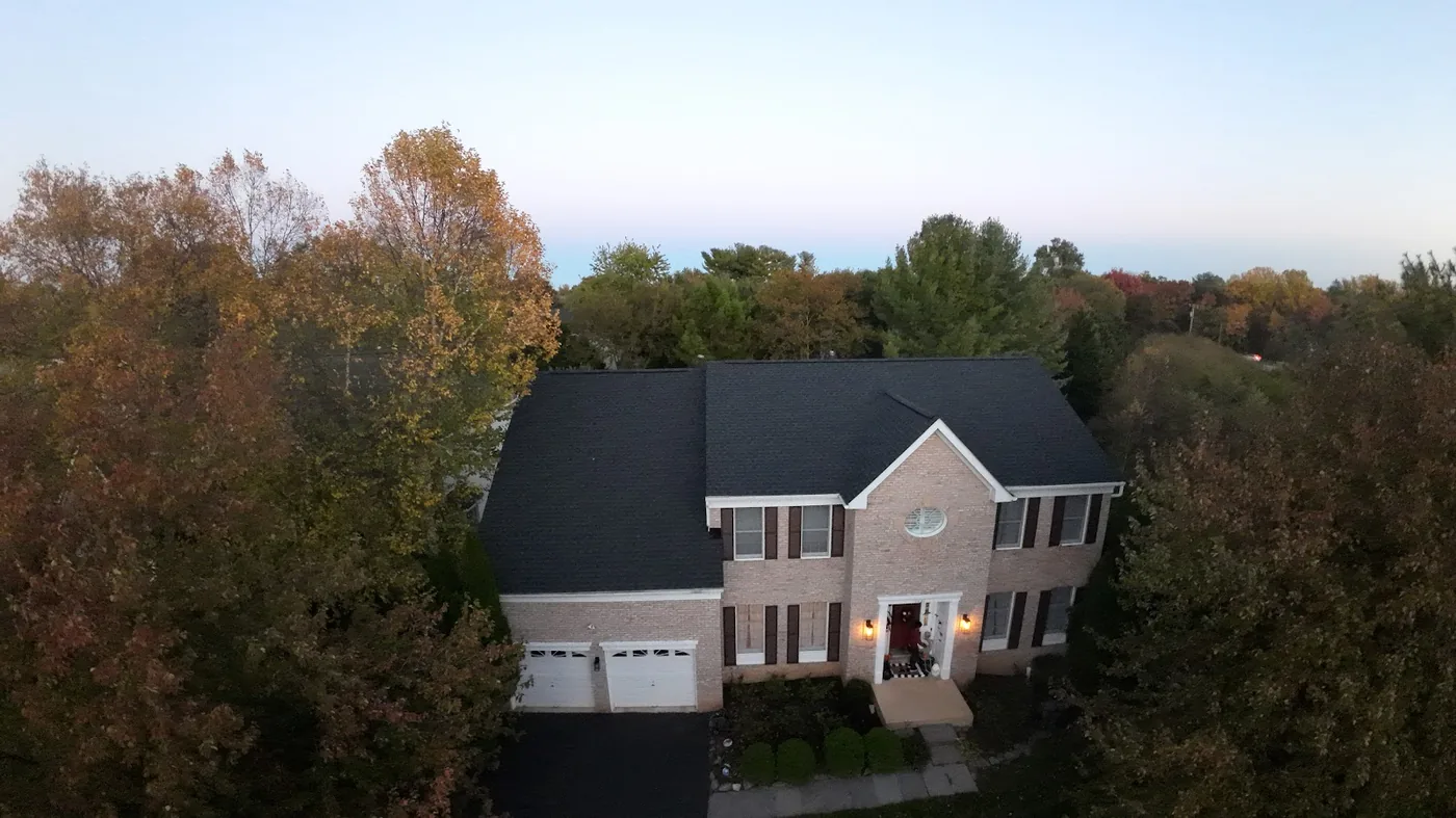Aerial view of a brick colonial house with a dark shingle roof surrounded by autumn trees