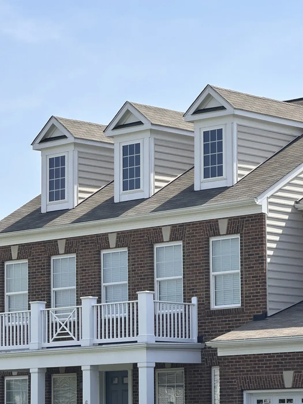 Three dormers on a brick house with a gray shingle roof and a white balcony railing